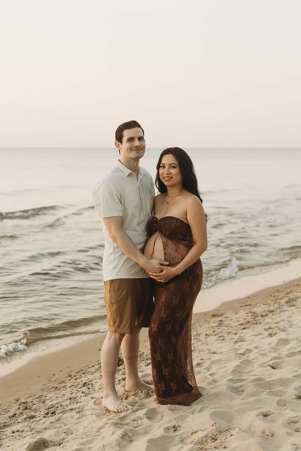A couple shares an intimate moment on a sandy beach at sunset as waves roll in along the shoreline.