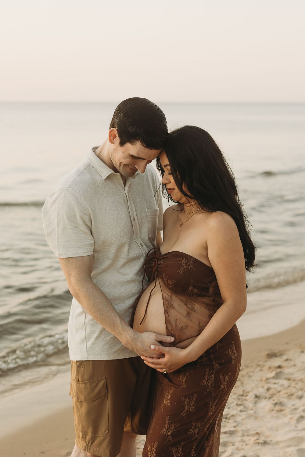 A couple shares an intimate moment together on a sandy beach at sunset with waves rolling in the background.