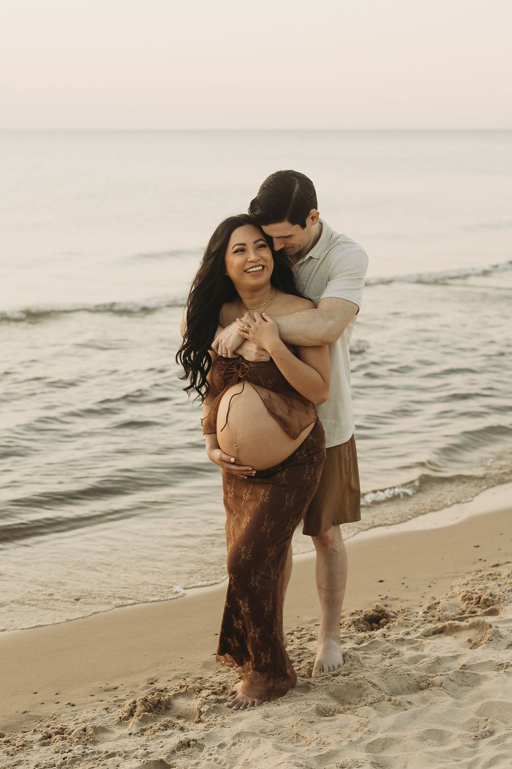 A couple embraces lovingly on a sandy beach at sunset with gentle waves rolling in the background.
