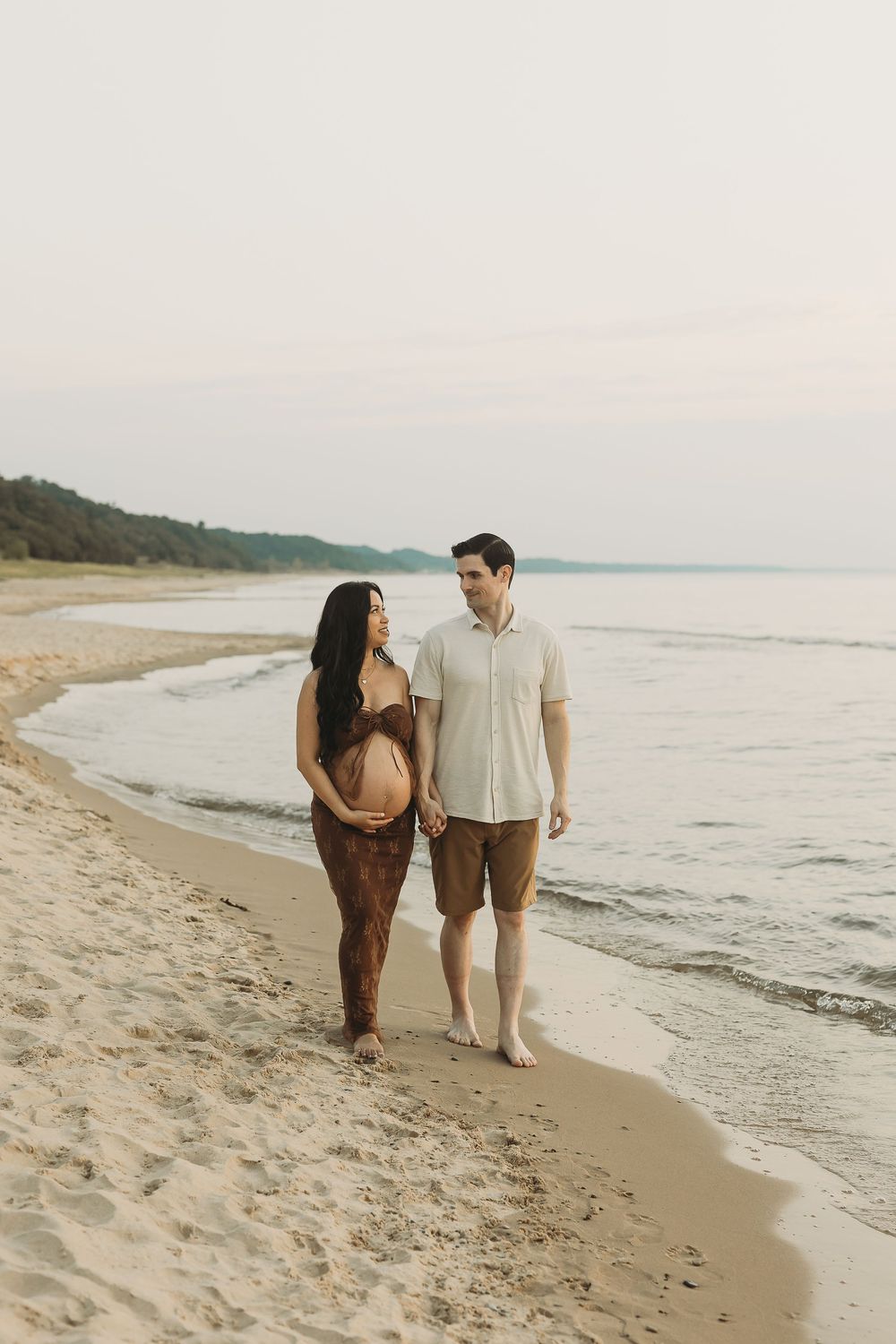 Two people stroll hand in hand on a peaceful beach with rolling waves and cloudy skies.