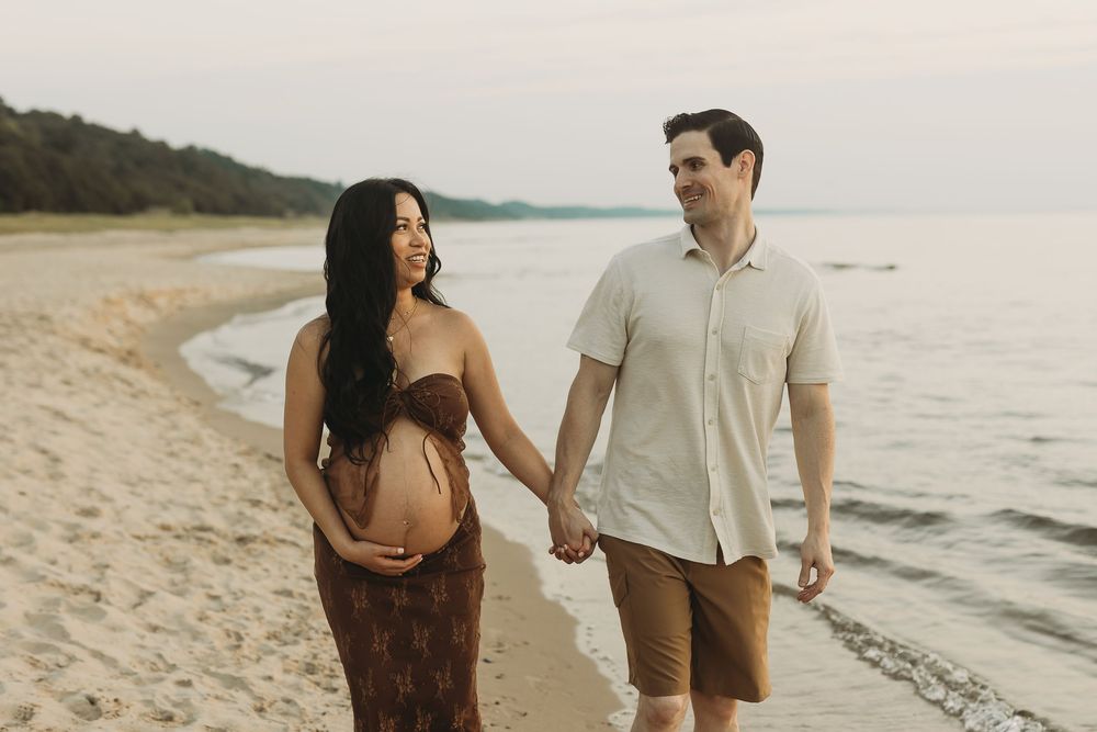 A couple walks hand in hand along a scenic beach at sunset wearing coordinated neutral-toned outfits.