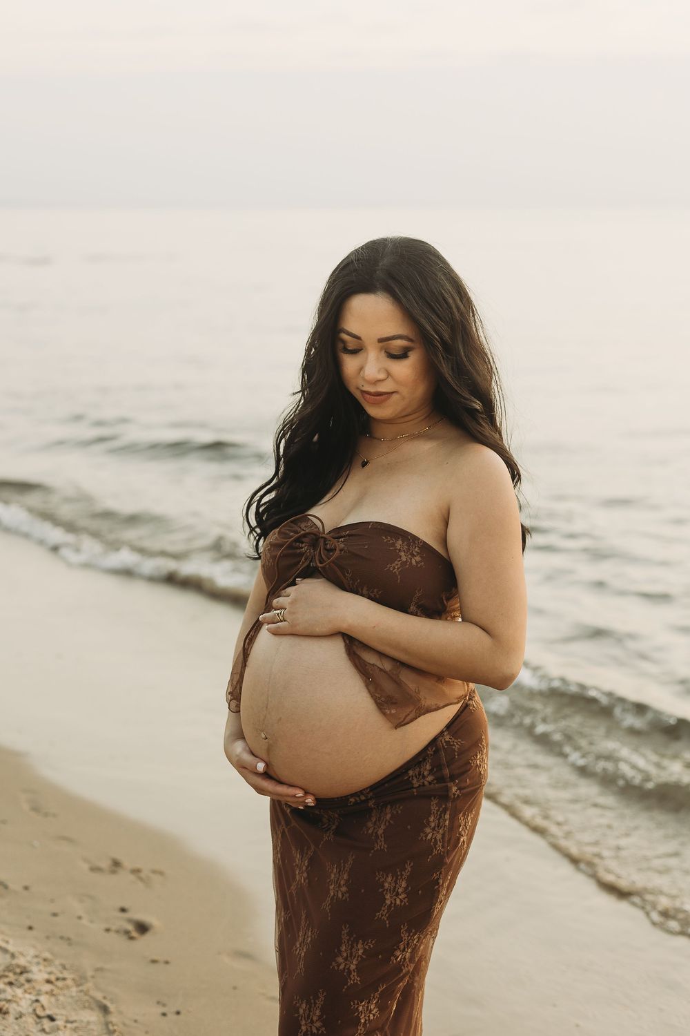 An expectant mother in a brown dress cradles her baby bump while standing on a sandy beach at dusk.
