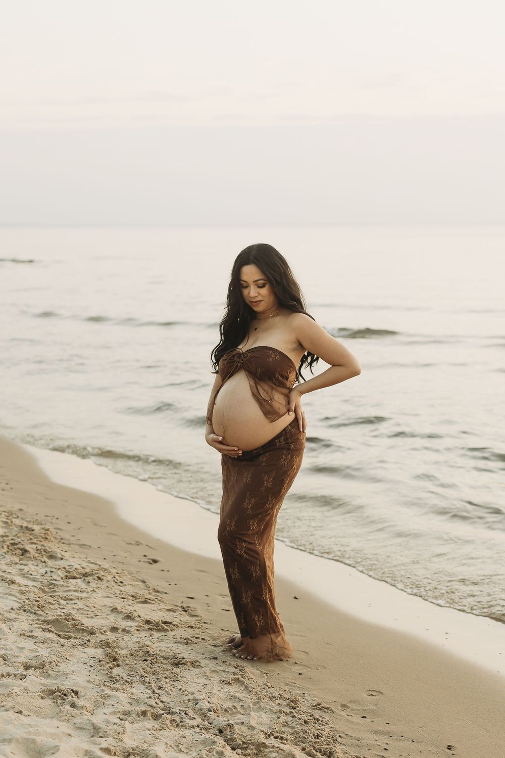 A pregnant woman in a brown dress poses gracefully on a misty beach at sunset for a maternity photoshoot.