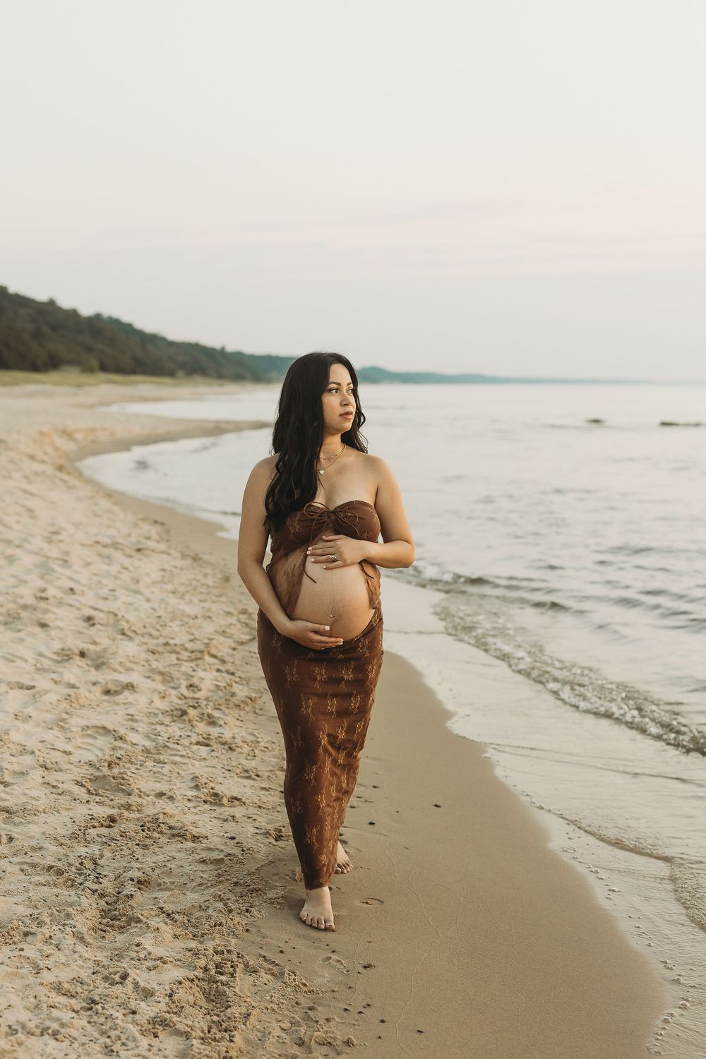 A maternity photo on a peaceful beach at sunset shows a pregnant woman in a brown dress walking along the shoreline.