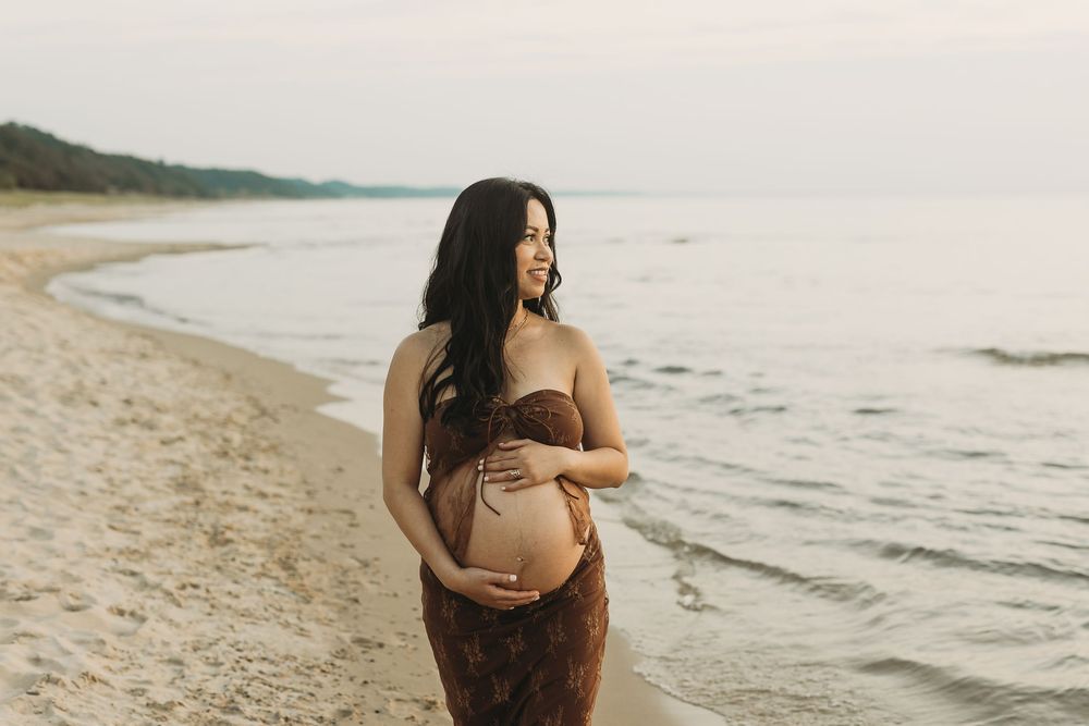 A pregnant woman in a brown dress walks along a scenic beach at sunset with waves gently rolling onto the shore.