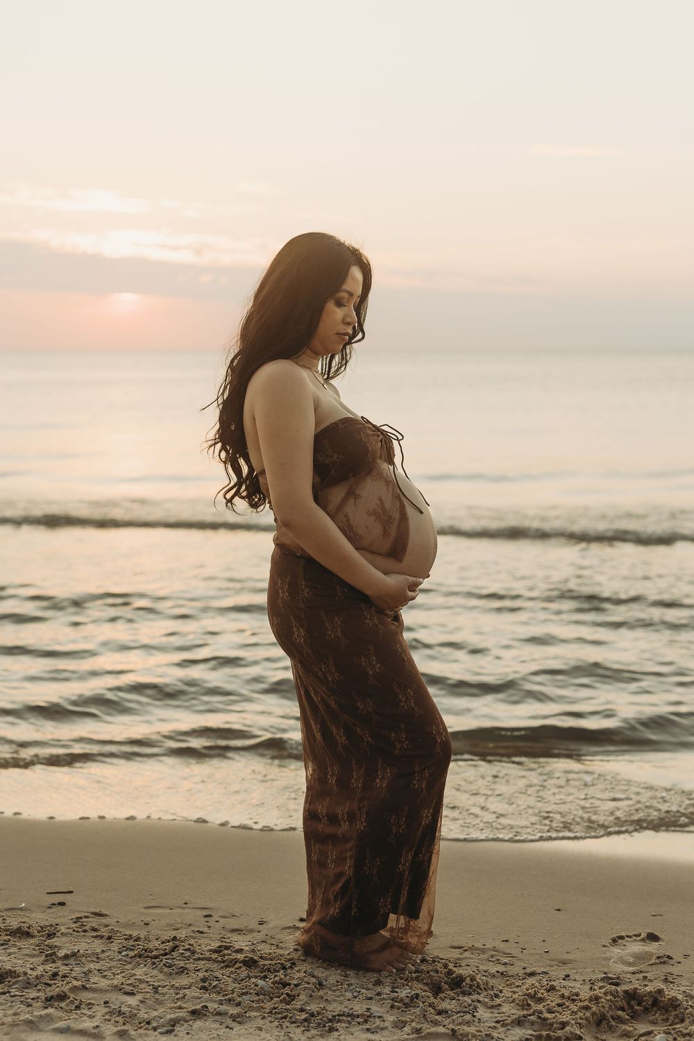 A couple embraces on a sandy beach at sunset with gentle waves in the background.