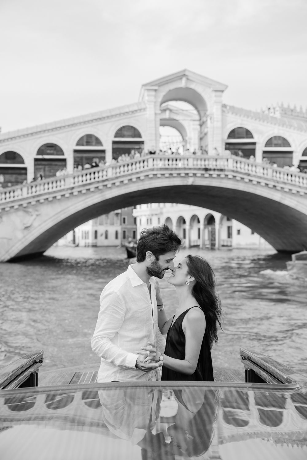 Black and white photo of a romantic couple kissing on a boat in front of the Rialto Bridge in Venice, Italy.