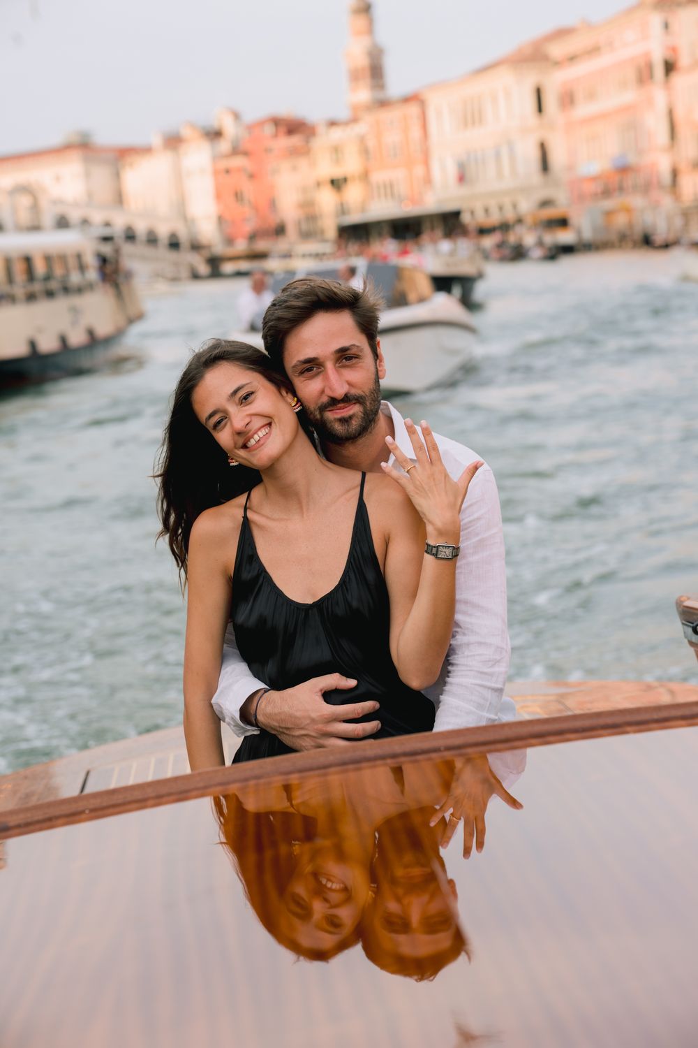 Happy couple posing on a boat in Venice, Italy, woman showing engagement ring with canal reflection.
