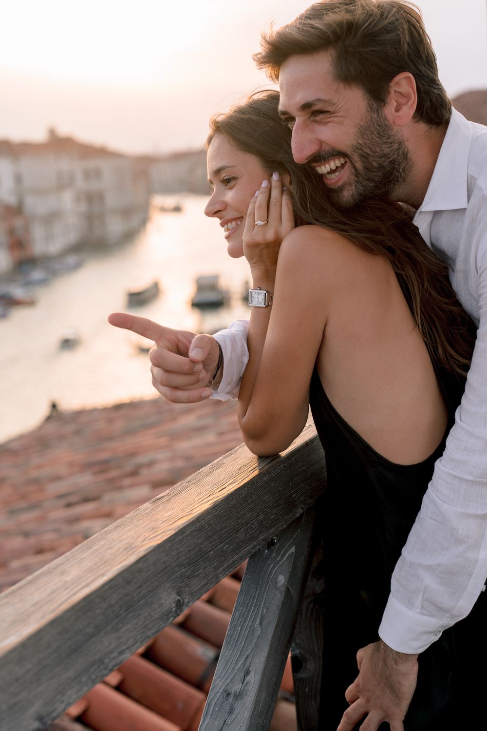 Happy couple on a Venice balcony at sunset, man pointing at Grand Canal view, woman in black dress smiling.