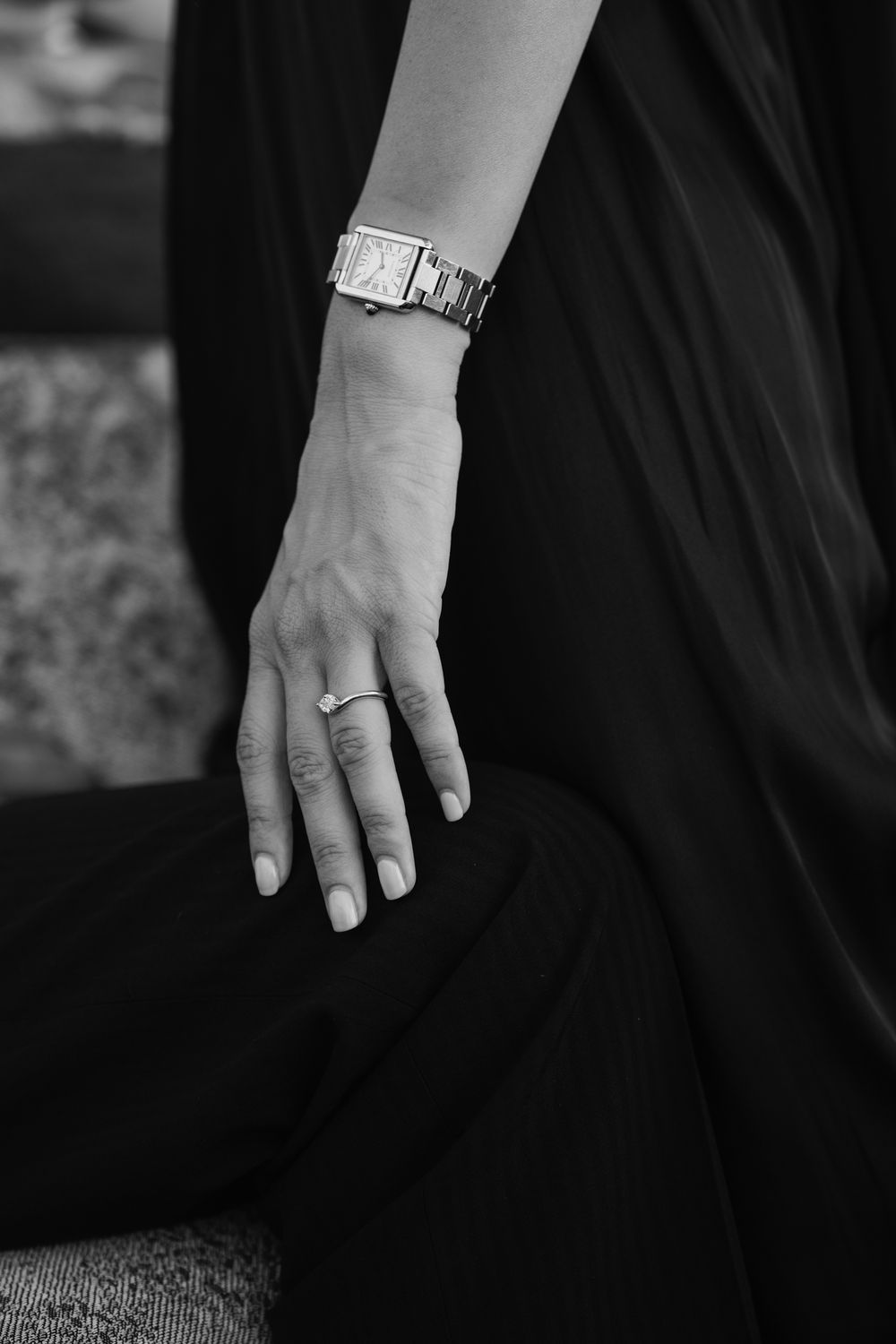 Black and white close-up of a woman's hand wearing a ring and elegant watch, resting on dark fabric.