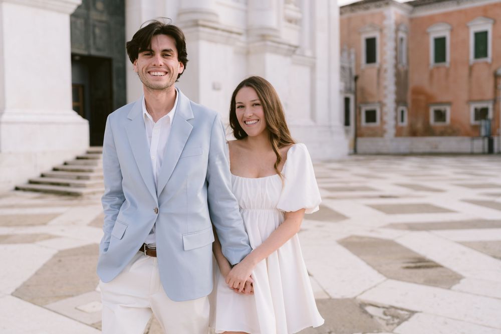 A couple in light summer attire holding hands and smiling in a picturesque Italian plaza with historic architecture.