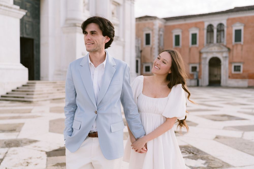 A couple in light blue suit and white dress hold hands while walking through an elegant Italian plaza.