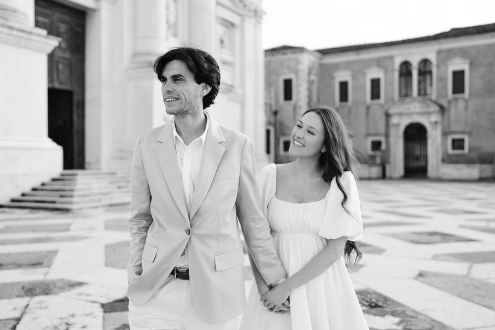 A black and white photo of a couple in light formal wear walking hand in hand through a European courtyard.
