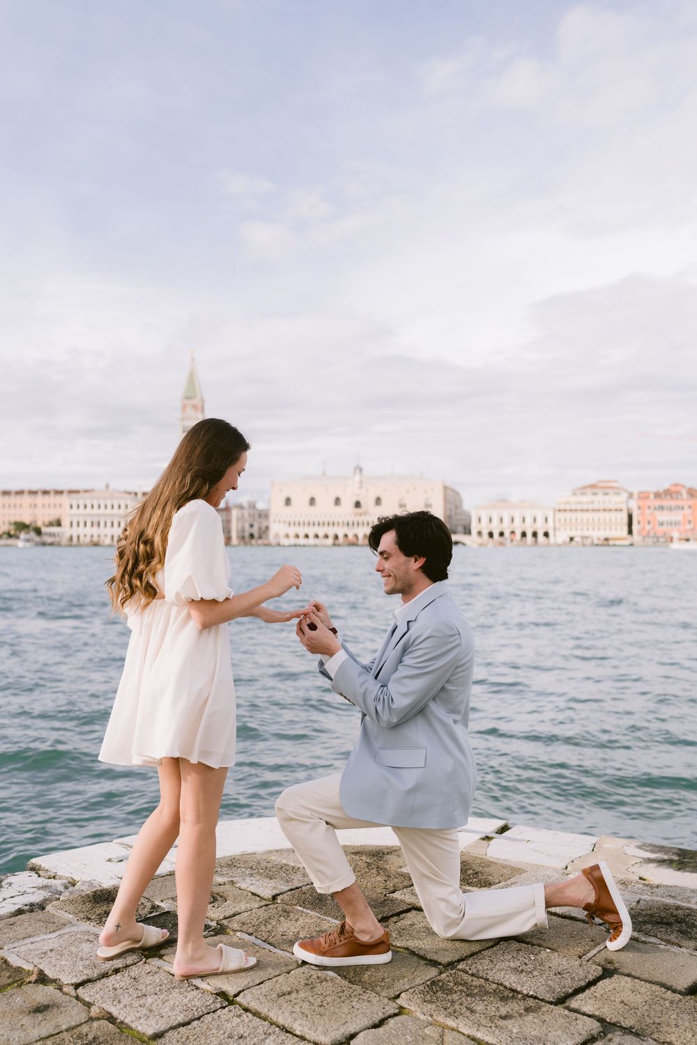 A romantic proposal scene unfolds along the waterfront in Venice with historic buildings visible across the lagoon.