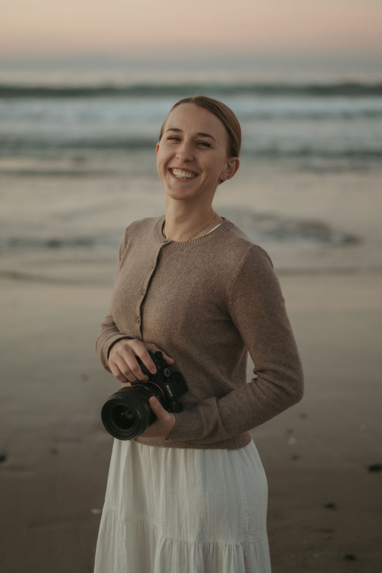 Person wearing brown sweater and white skirt poses casually on beach at sunset with ocean waves in background.