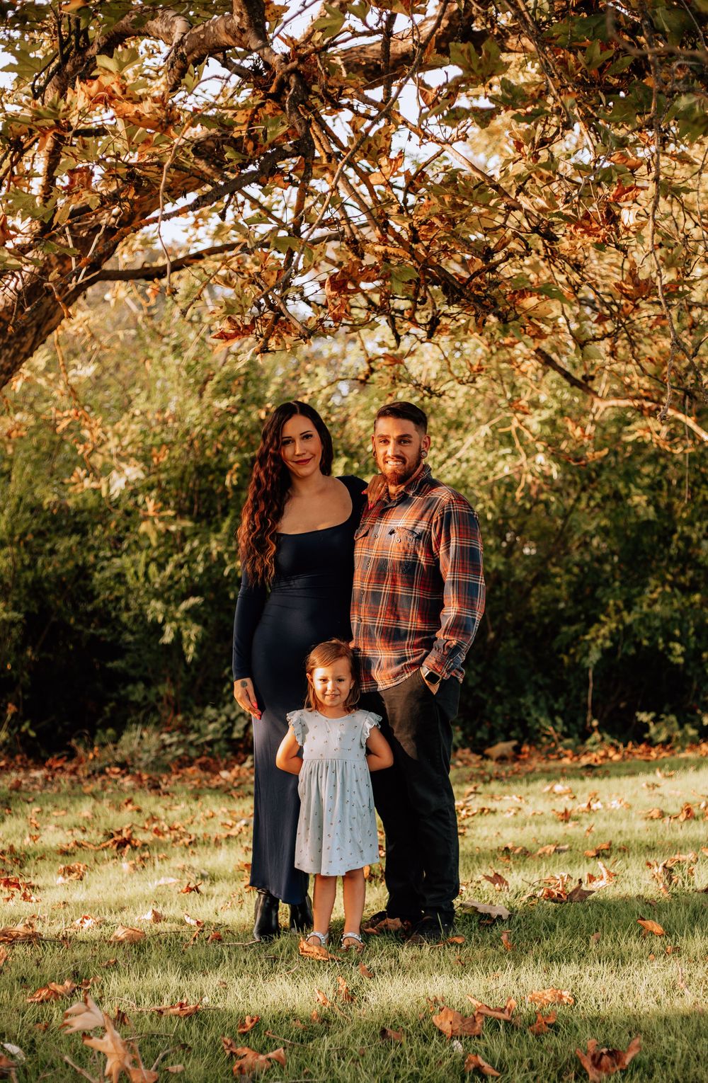 A family stands together under autumn trees with golden leaves on a sunny afternoon in a park setting.