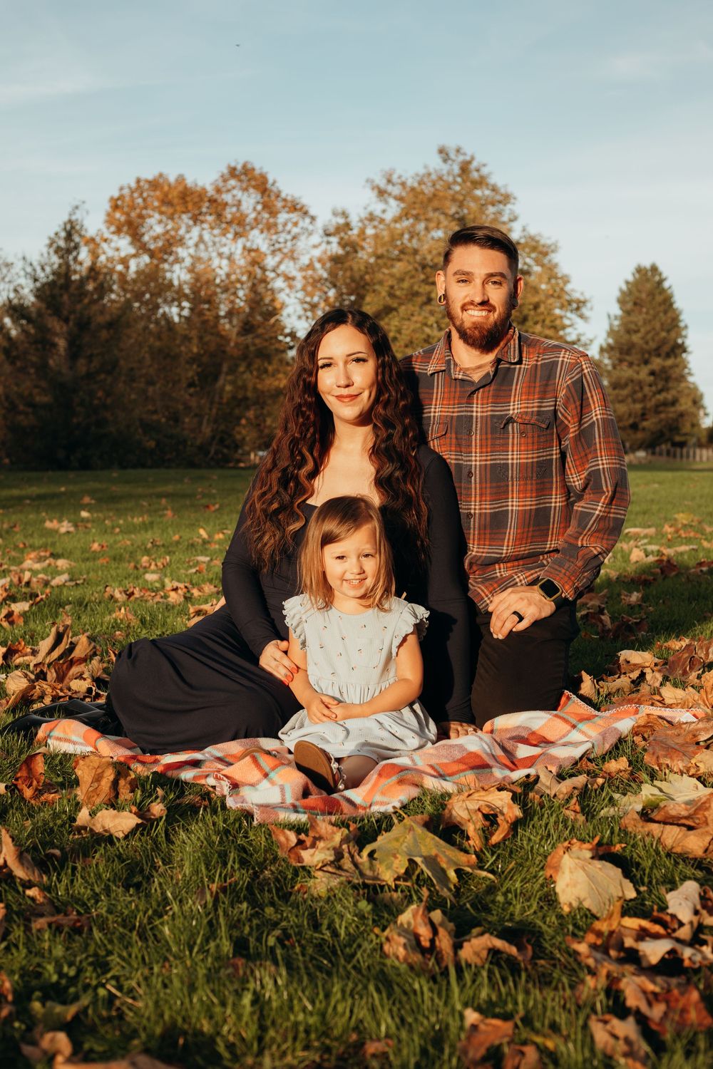 Family relaxes on a blanket surrounded by fallen autumn leaves during a golden sunset photoshoot.