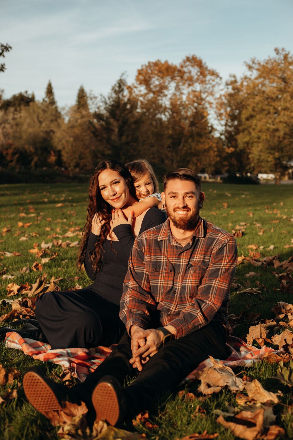 A couple relaxes on grass surrounded by fallen autumn leaves during sunset in a park.