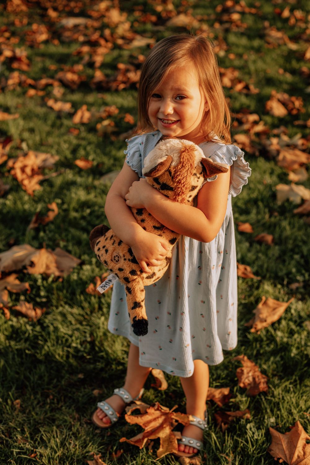 A child in a white dress plays with autumn leaves on grass during golden hour sunset lighting.