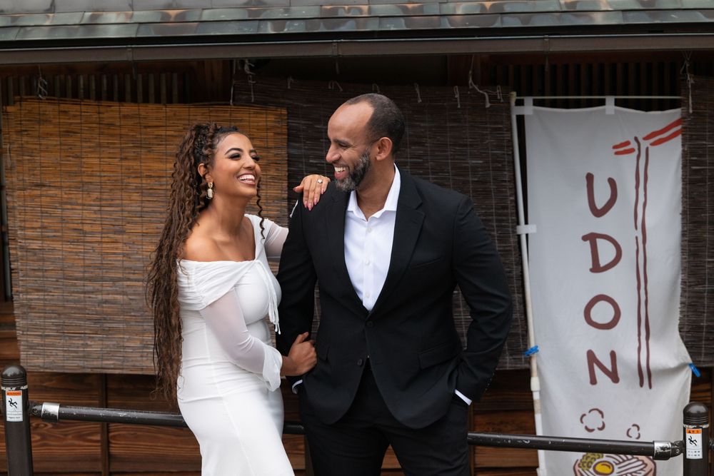 Smiling couple stands outside Japanese udon restaurant, woman in white dress, man in black suit.