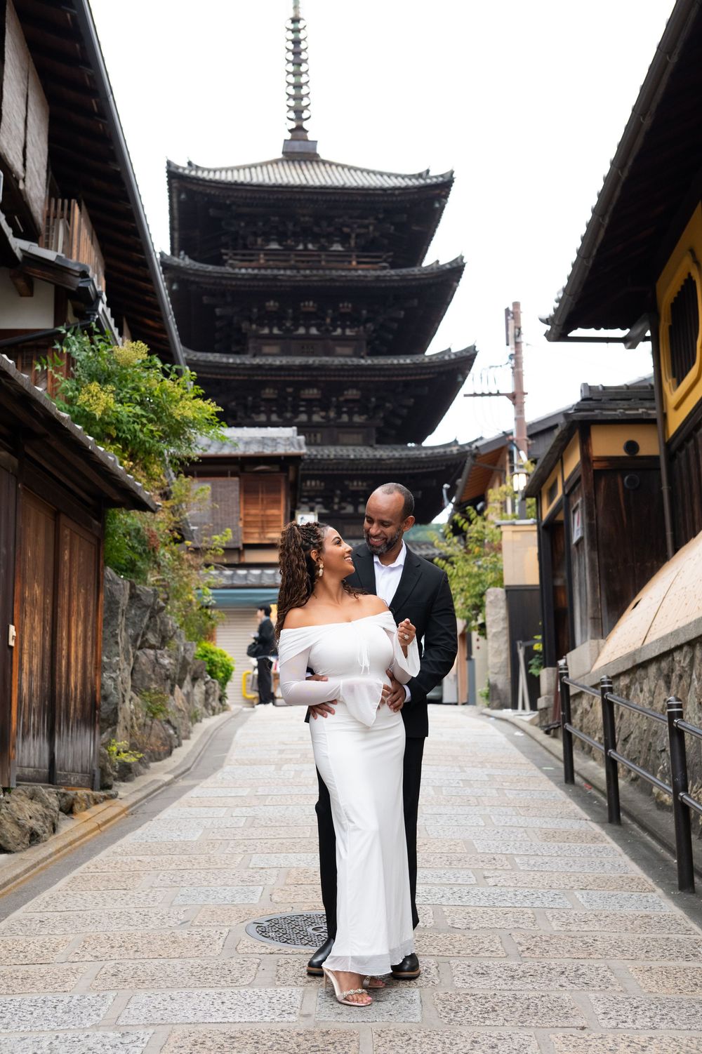 Couple embraces on Kyoto stone path with Yasaka Pagoda behind, woman in white dress, Japan.