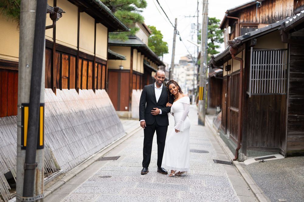 Couple walks hand-in-hand down a traditional Japanese street in Kyoto, woman in white dress, man in black suit.