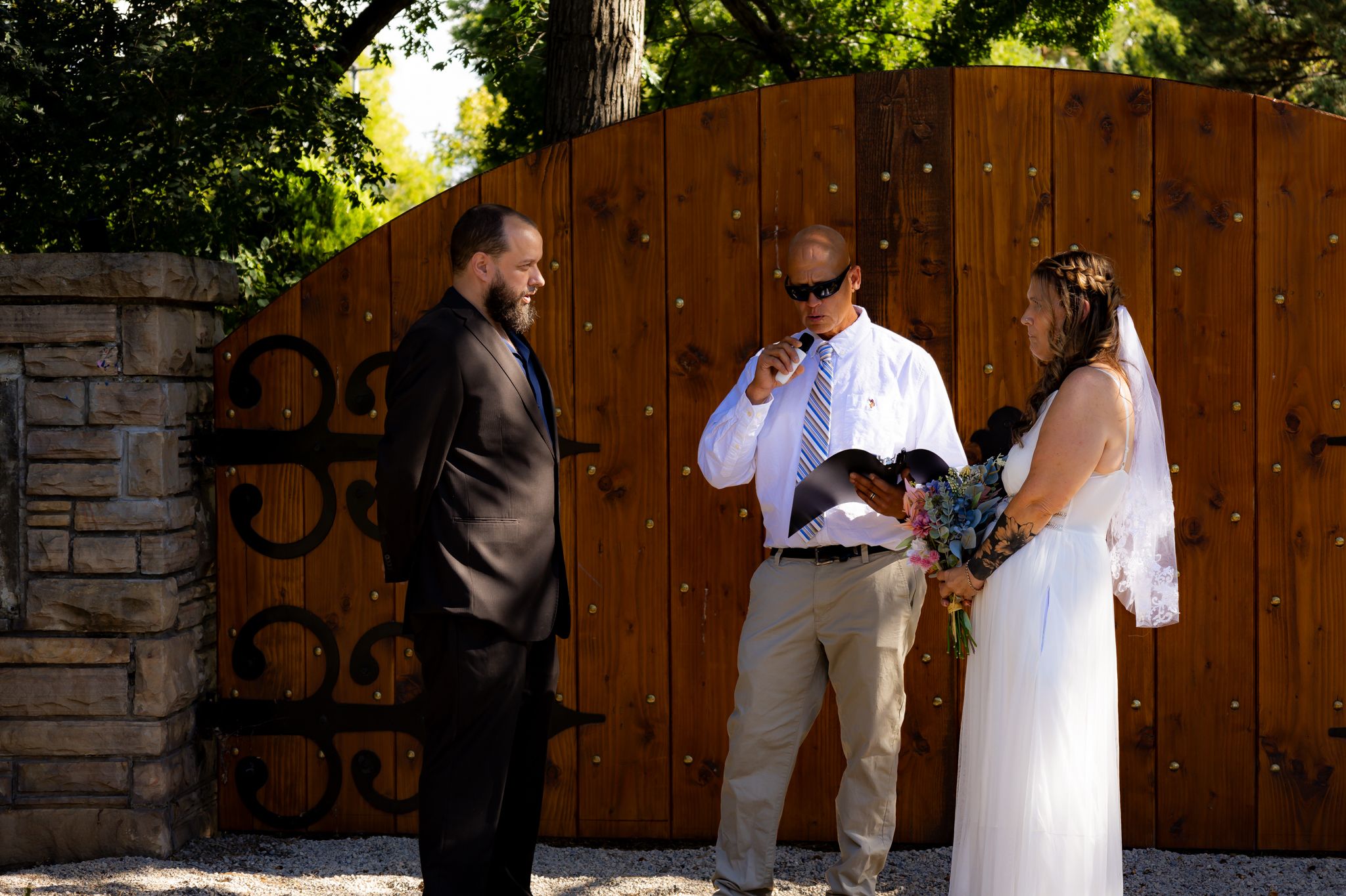 An outdoor wedding ceremony with participants gathered in front of a rustic wooden gate.