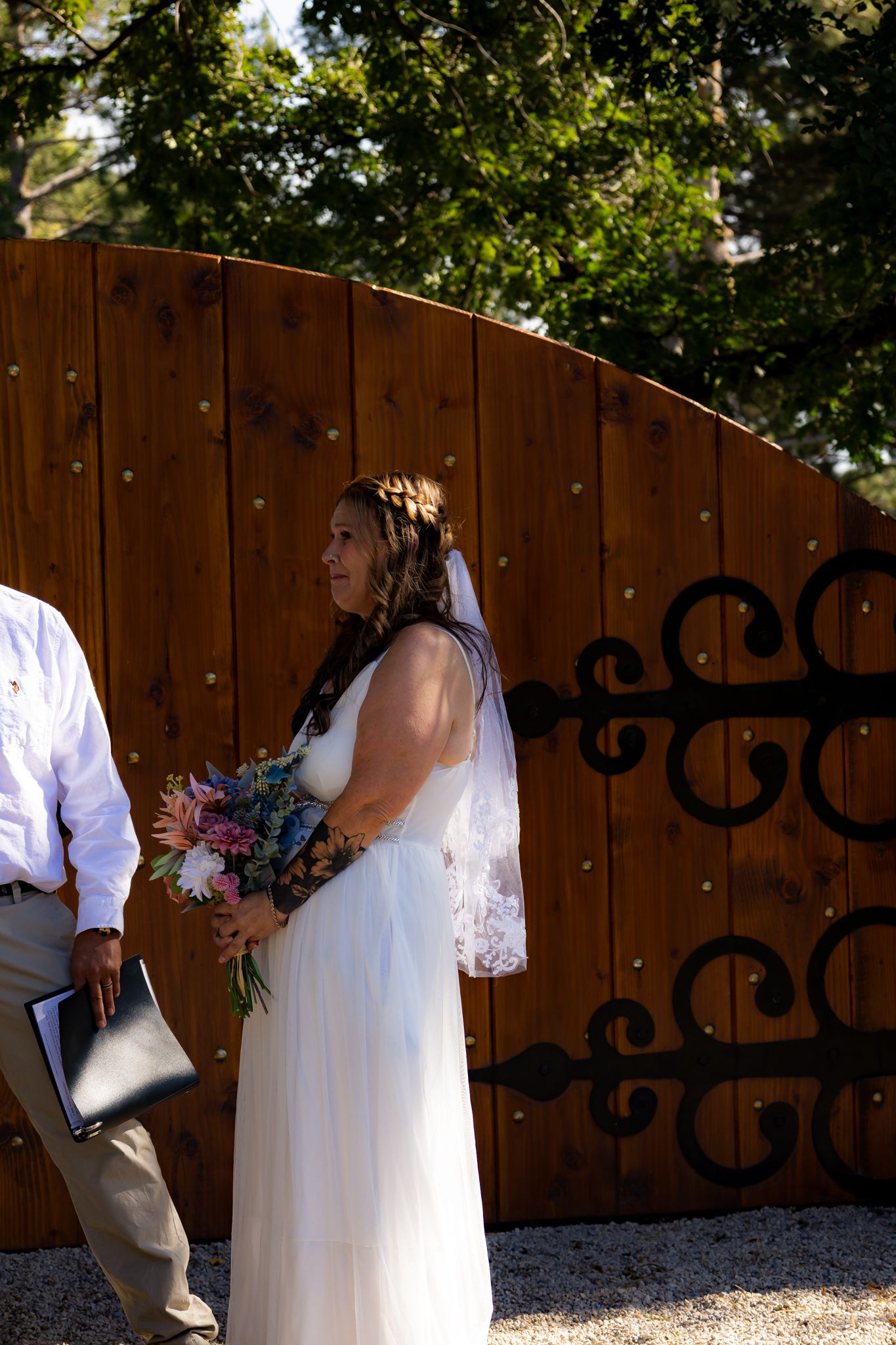 A wedding ceremony participant holding a colorful bouquet stands against a wooden gate backdrop.
