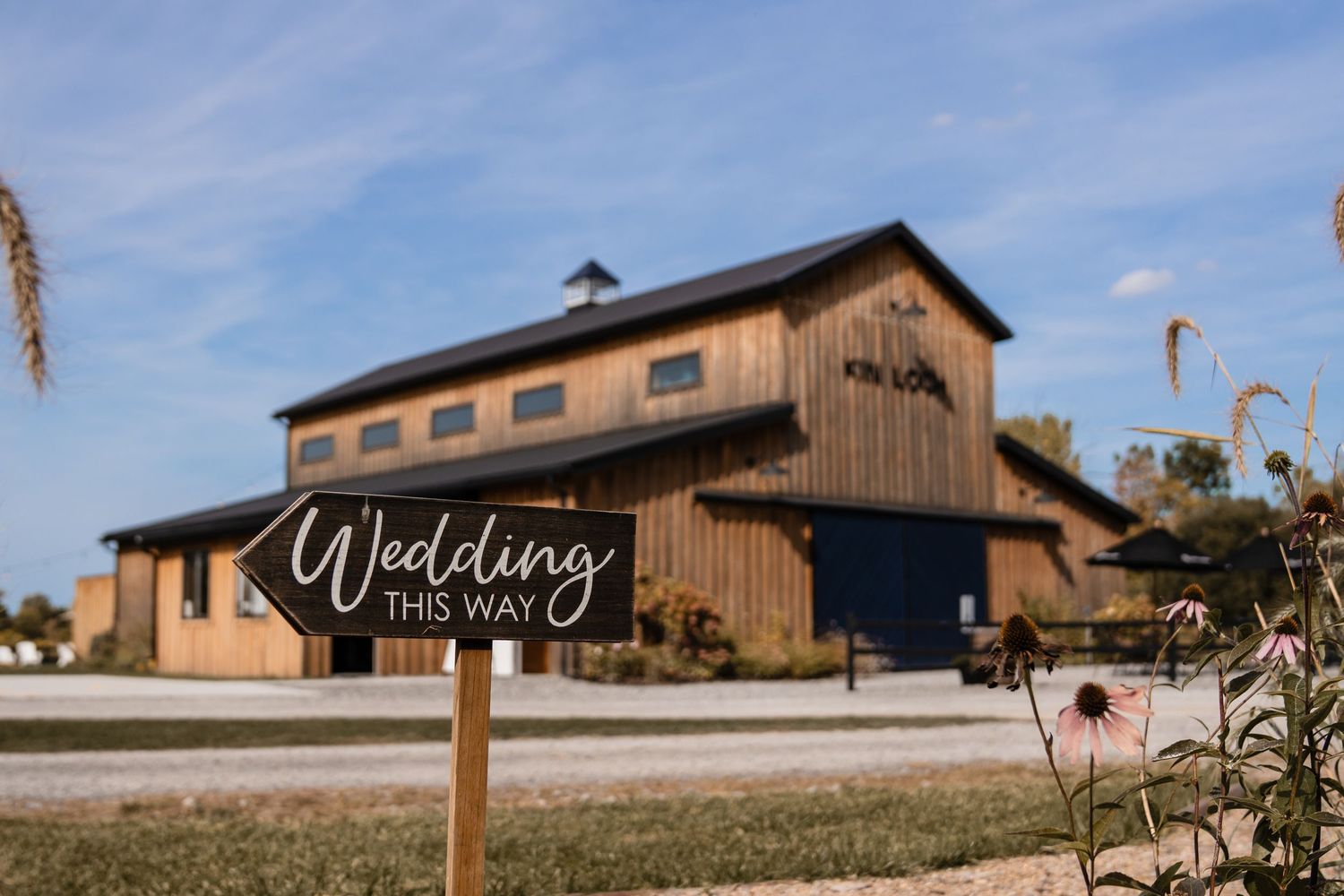 A wedding ceremony takes place outdoors under willow trees with guests seated in white chairs.
