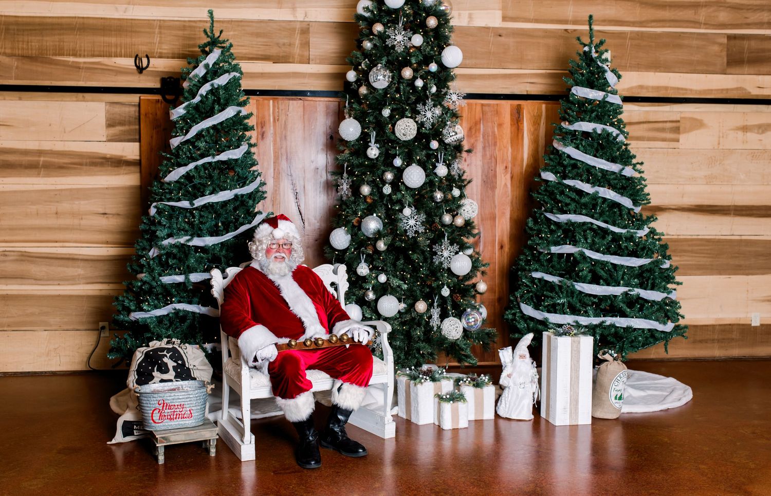 A series of Christmas trees and holiday decorations arranged against a wooden wall with festive lighting and presents.