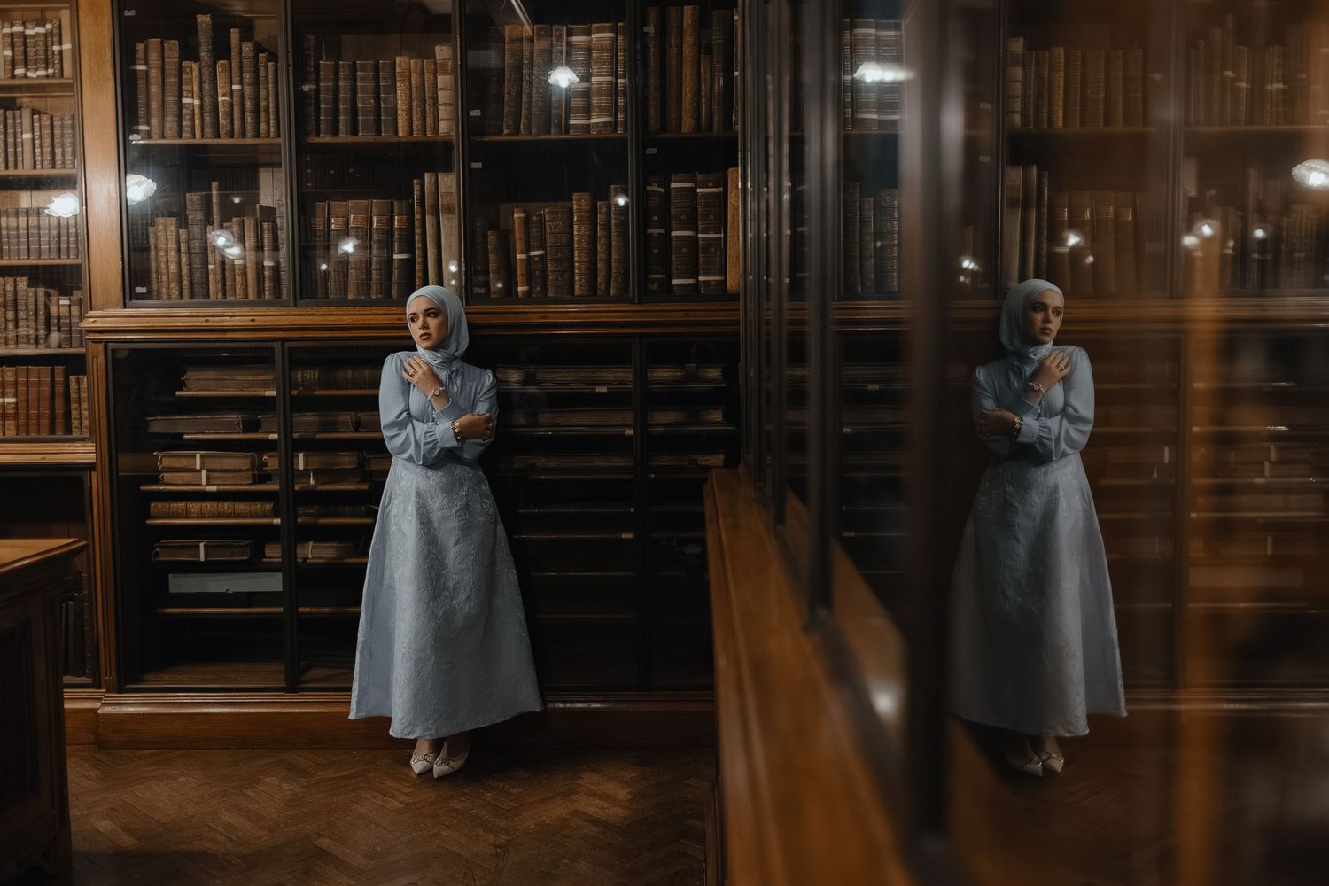 A person in a light gray hijab stands in front of wooden library shelves filled with vintage books.