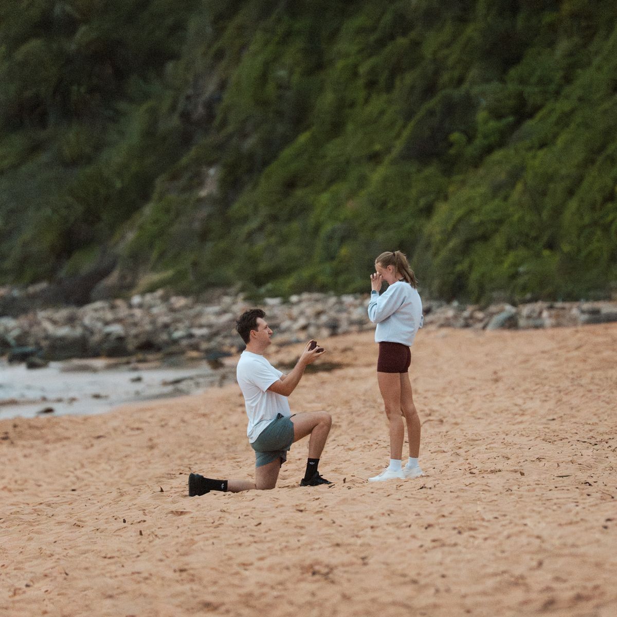 A proposal scene in sequence as a couple stands on a scenic beach with coastal cliffs in the background in newport photoshoot in Sydney