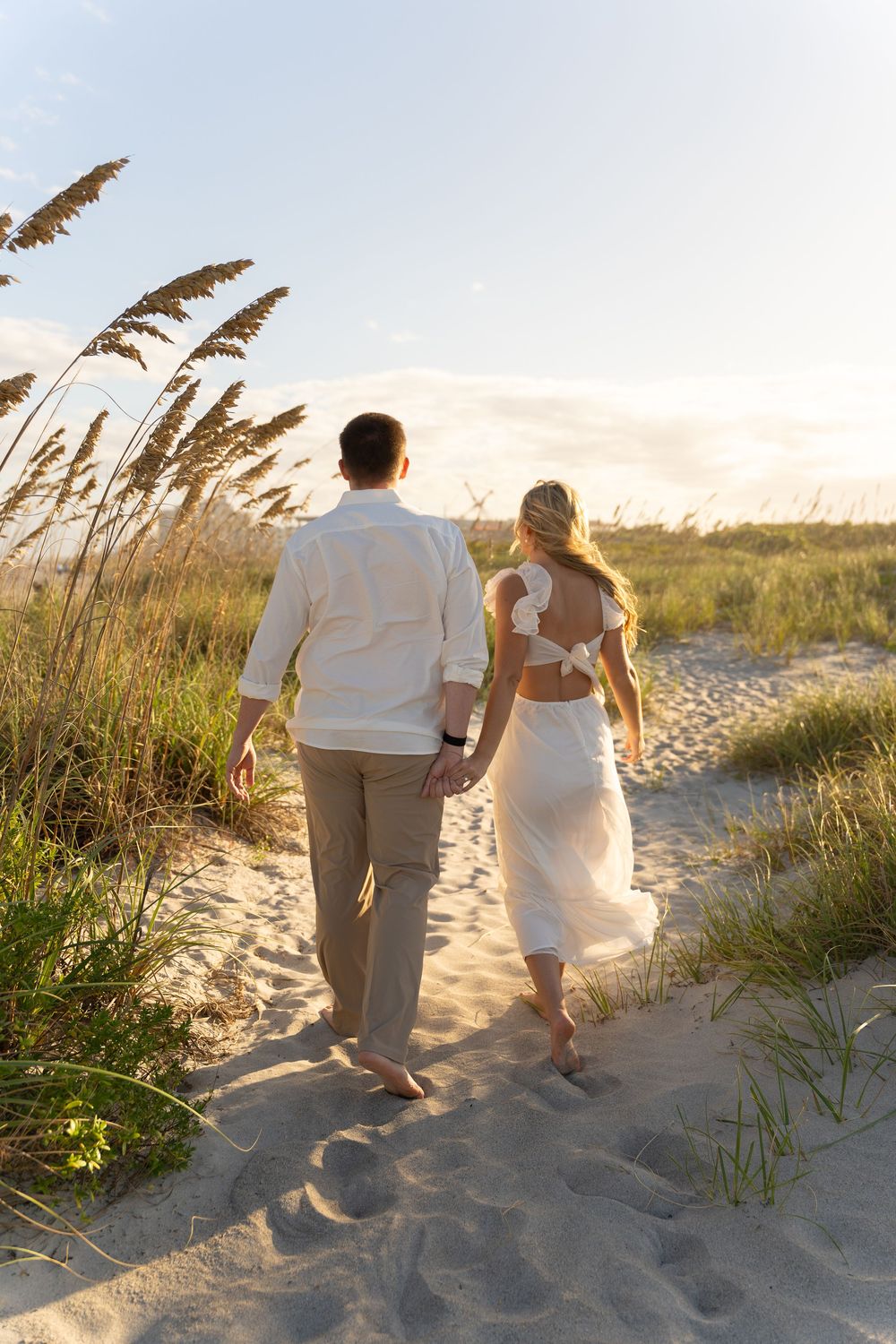 A couple in white attire walking together on a sandy beach path through sea oats at sunset.