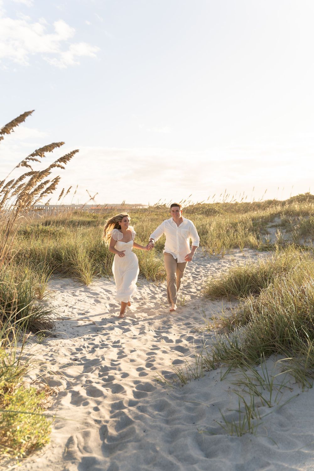 Couple running playfully together on a winding beach path through coastal dunes.