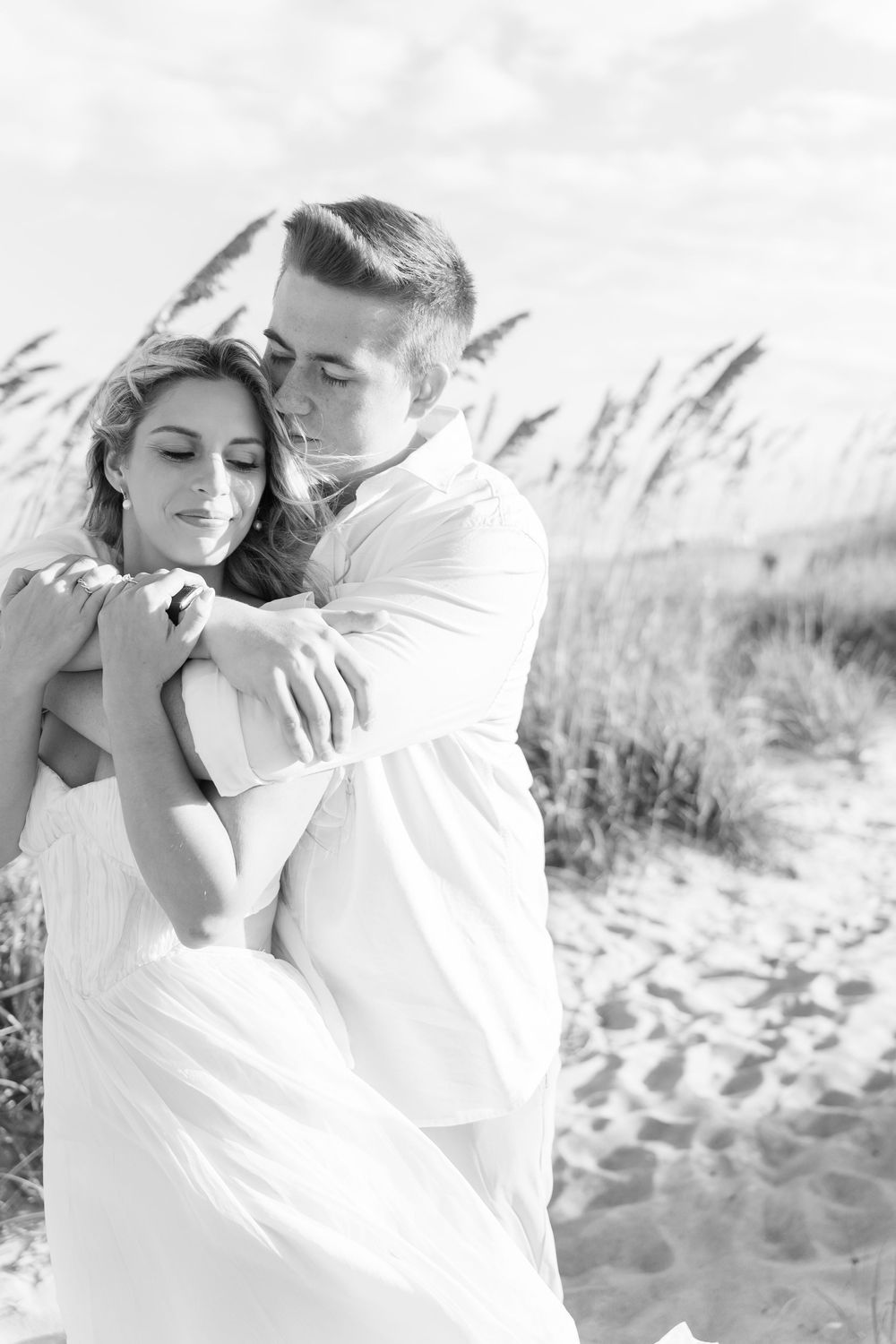Romantic black and white beach photos of a couple embracing against sand dunes and sea grass.