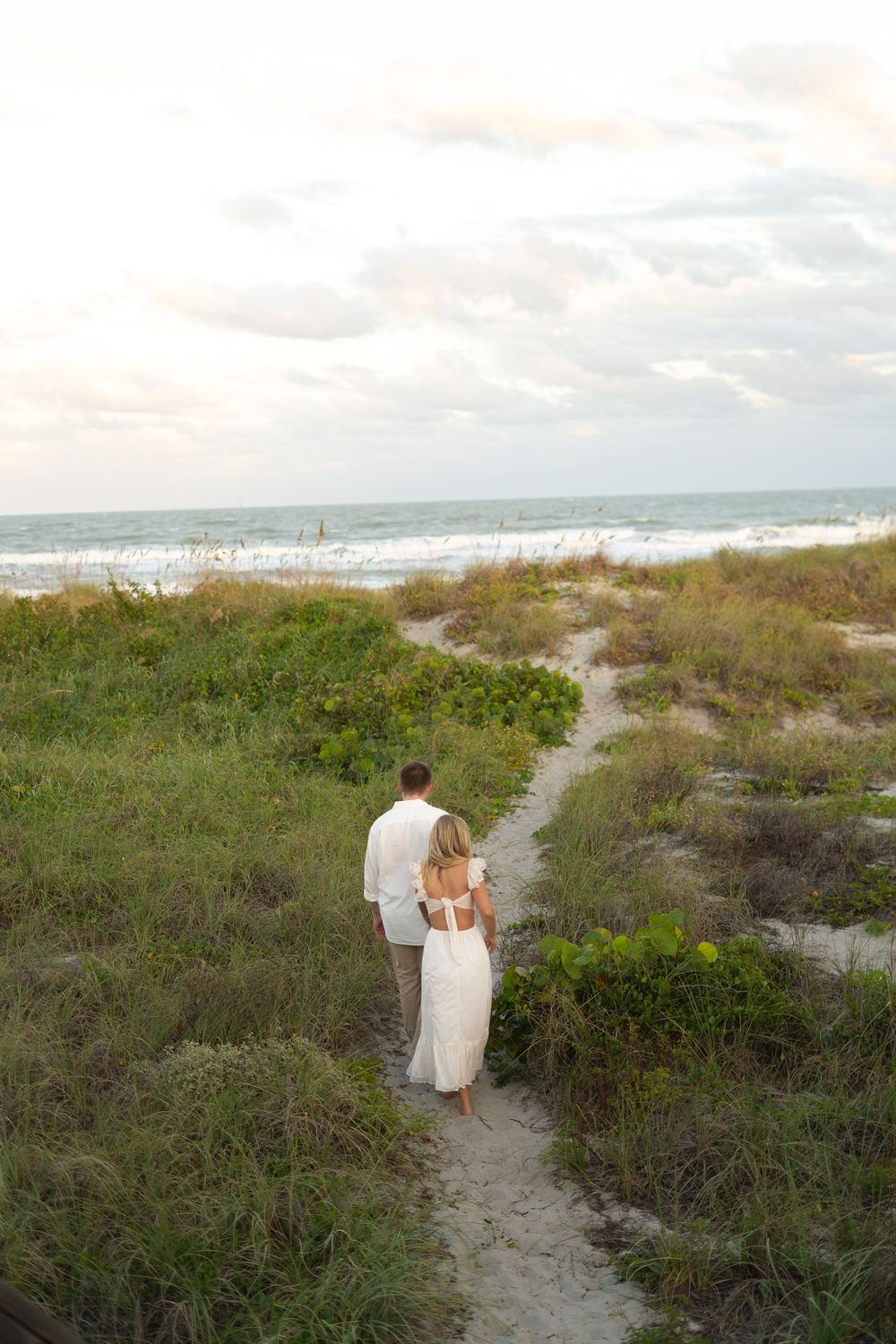 A couple walks along a coastal path through sand dunes and beach vegetation.