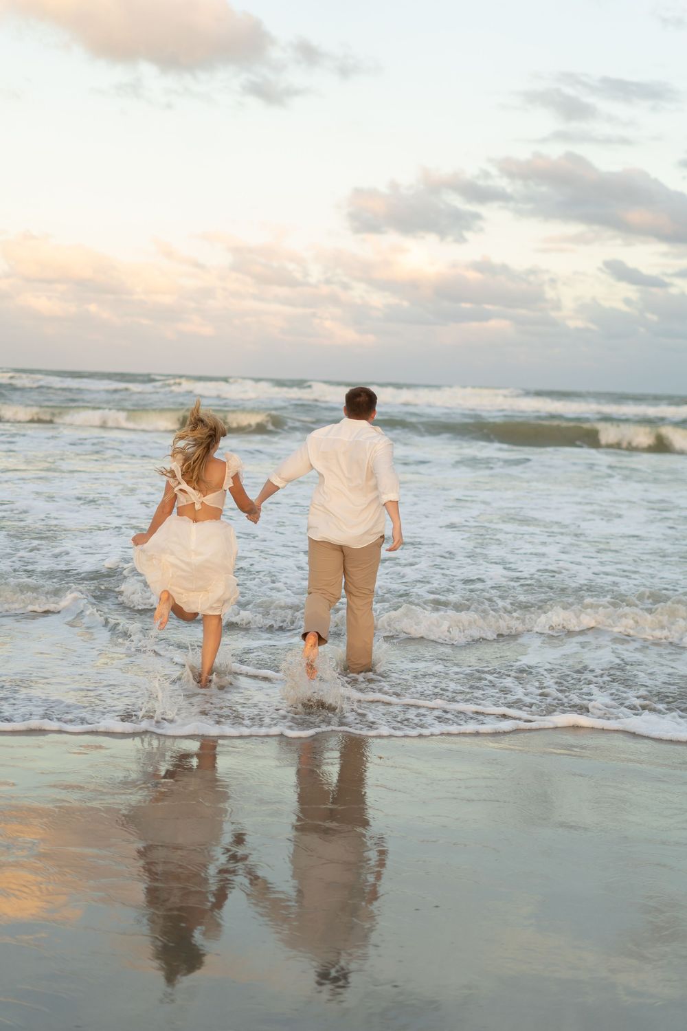 A couple playfully runs and splashes in the ocean waves at sunset on a sandy beach.