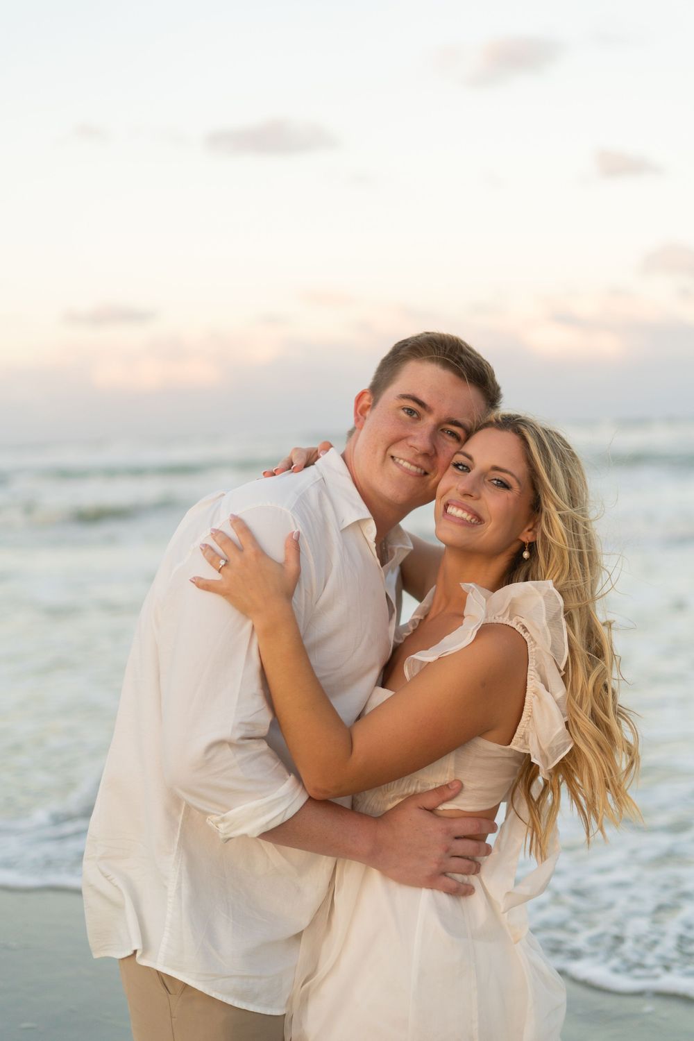 A couple in white outfits embrace and laugh together on a beach at sunset with waves in the background.