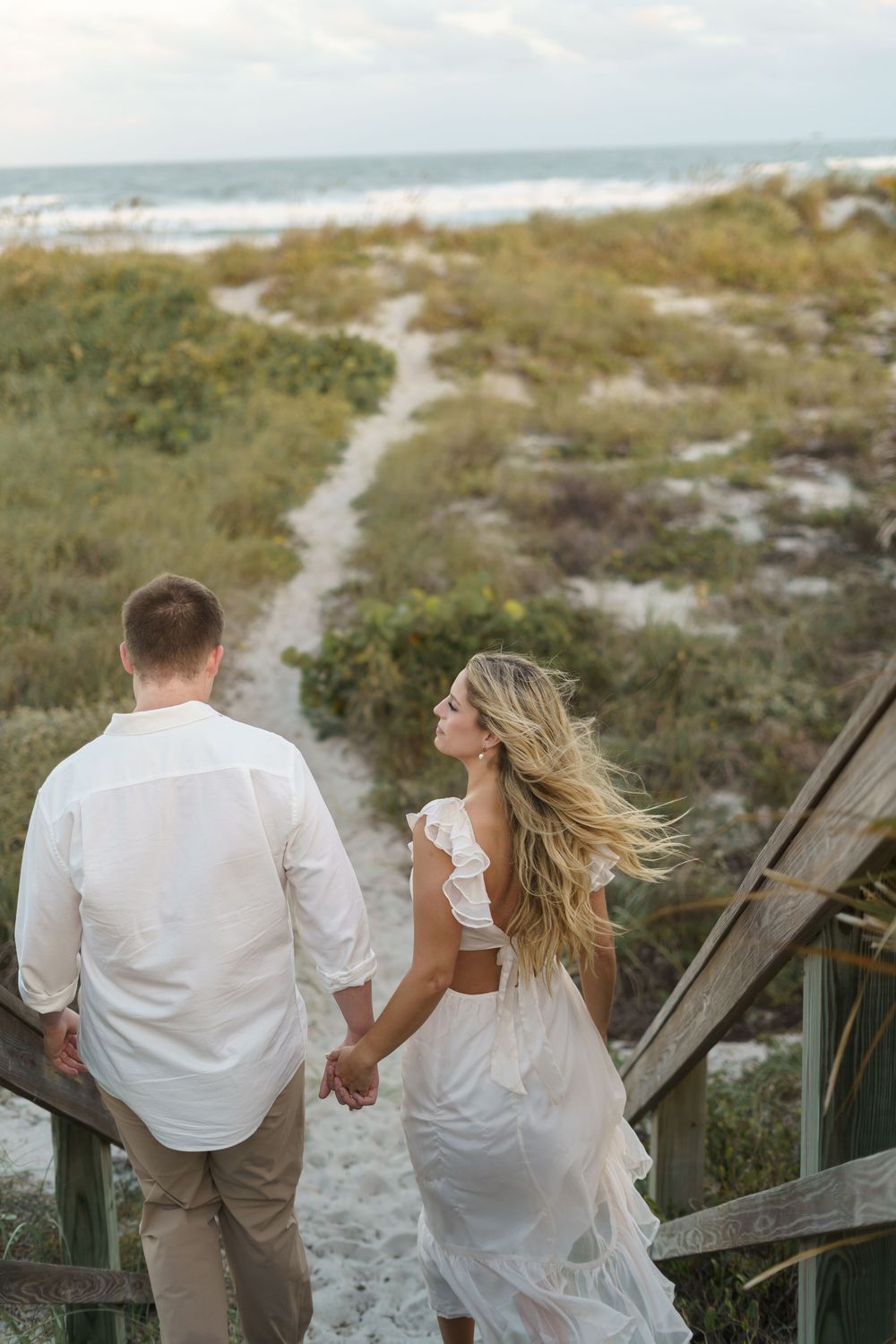 Couple walking hand in hand along a sandy beach boardwalk surrounded by coastal vegetation at sunset.
