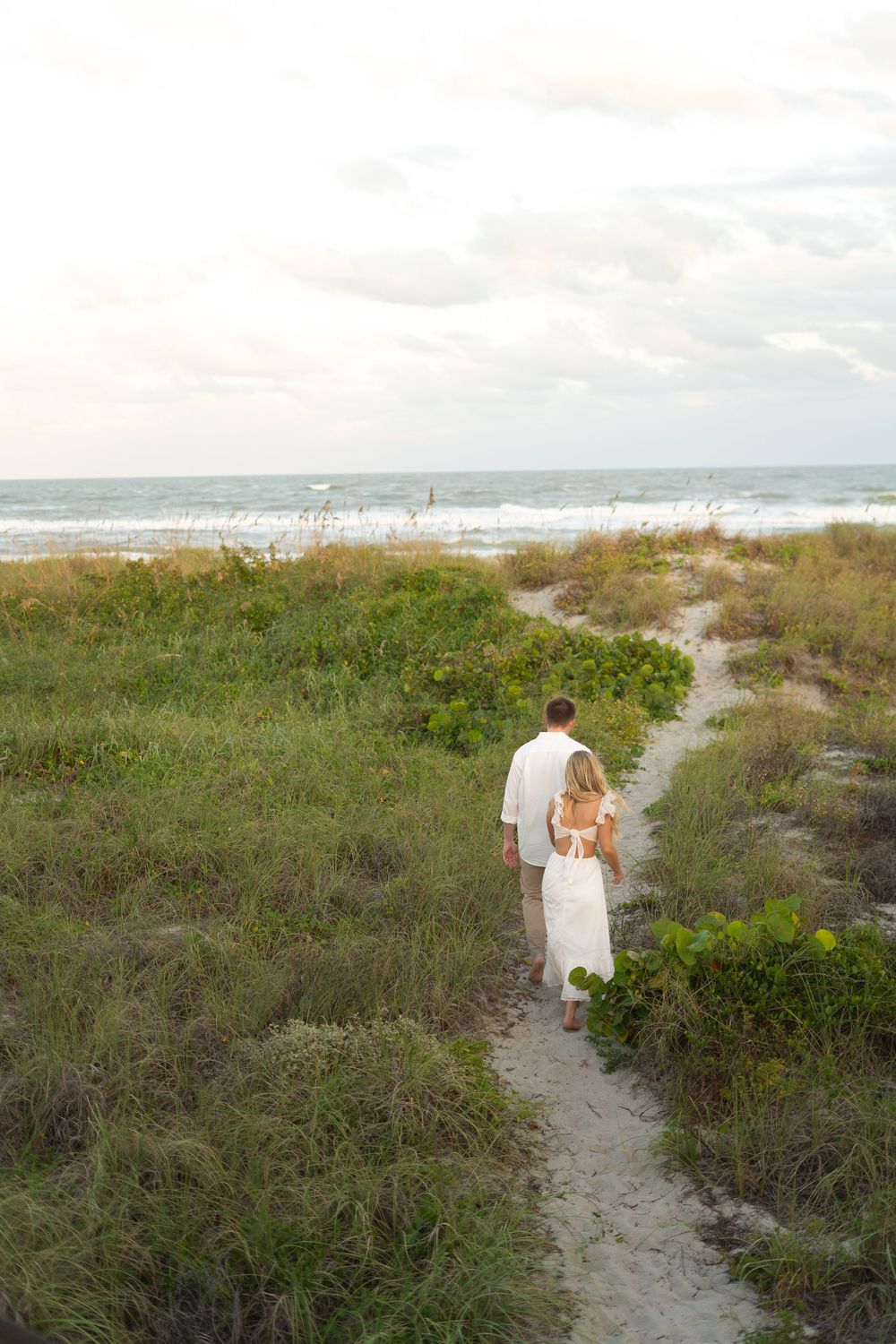Two people walk hand in hand down a sandy path through coastal vegetation with ocean views.