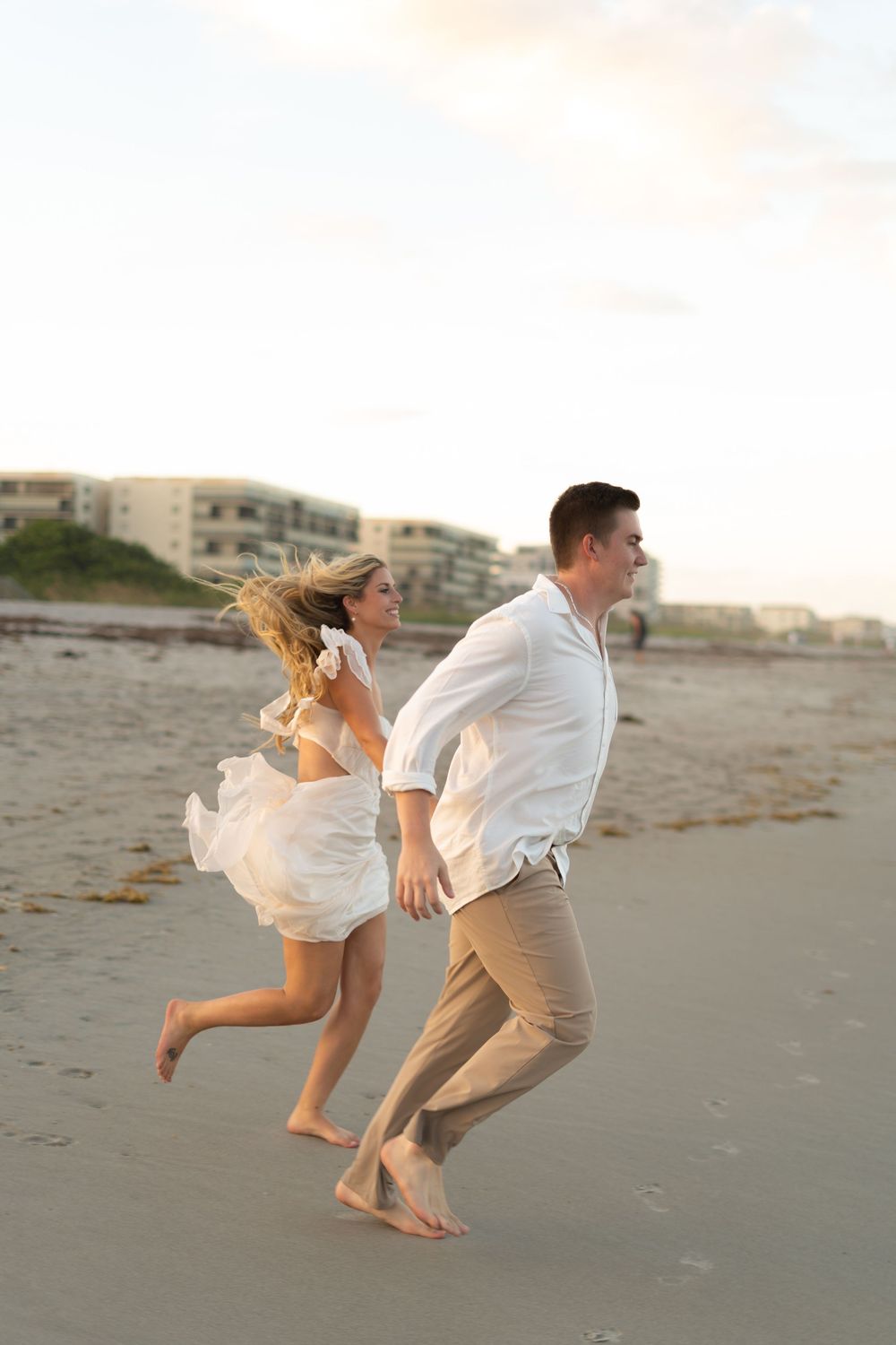 A playful couple in white clothing runs and dances together on a sandy beach at dusk.