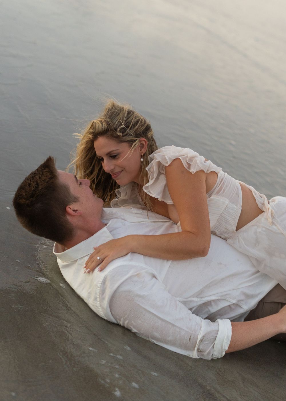 Wedding couple shares playful moments lying in shallow ocean water wearing white attire.