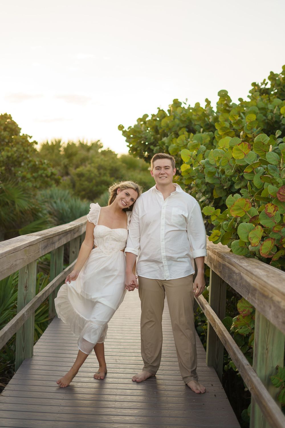 Couple in white and beige outfits posing playfully on a wooden boardwalk surrounded by sea grape plants.