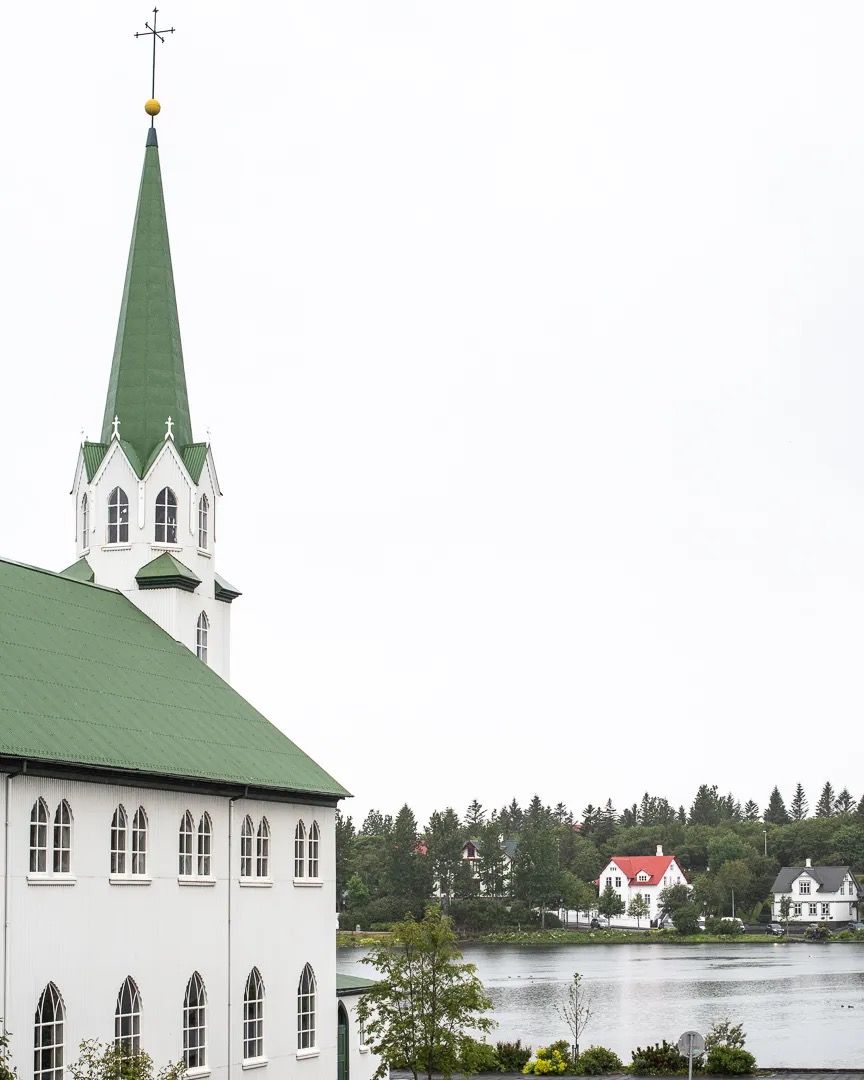 White church building with a tall green steeple overlooking water.