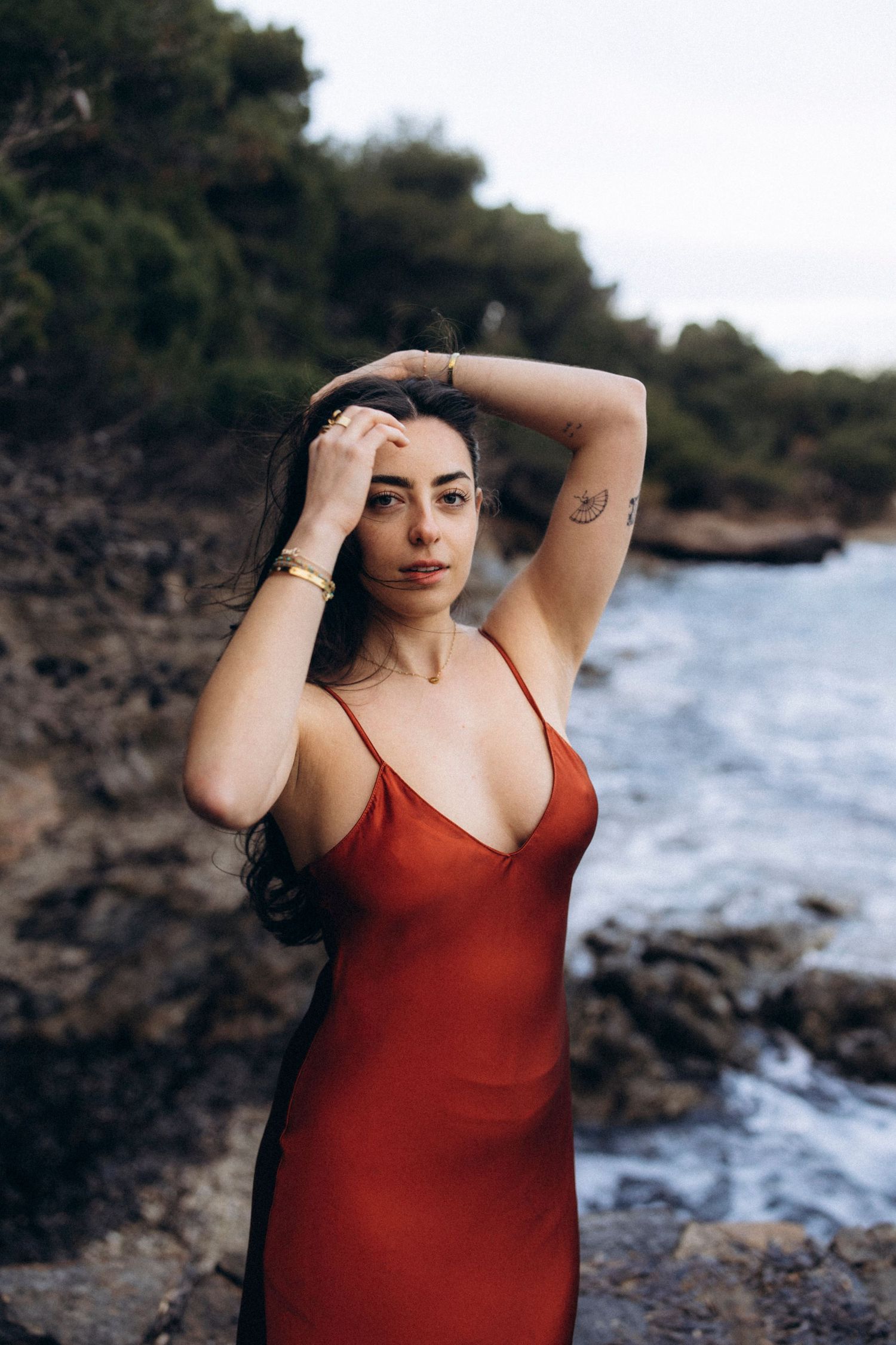 Person in a red dress poses near rocky coastline with ocean waves and dark foliage in background.