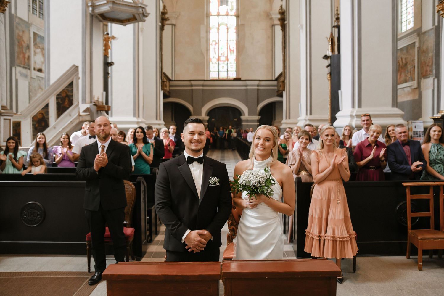 Church wedding ceremony showing guests seated in wooden pews with sunlight streaming through stained glass windows.