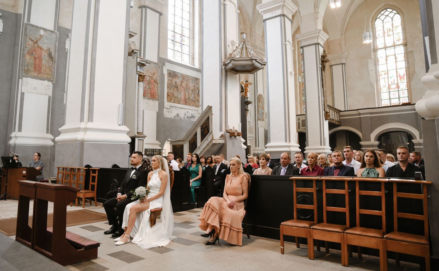 A panoramic view of a traditional church interior with wooden pews and high arched windows during a wedding ceremony.