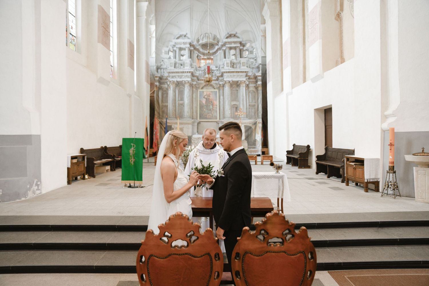 Wedding ceremony takes place in a bright, ornate church sanctuary with white columns and decorative altar.
