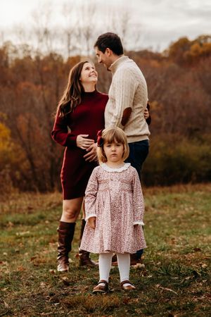 A young child in a pink floral dress stands in a grassy autumn field while adults embrace in the background.