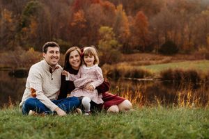A family sits together in a grassy meadow surrounded by autumn foliage during a golden hour photoshoot.